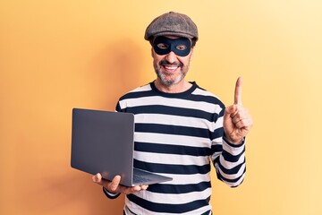Middle age handsome burglar man wearing cap and mask using laptop over yellow background smiling with an idea or question pointing finger with happy face, number one