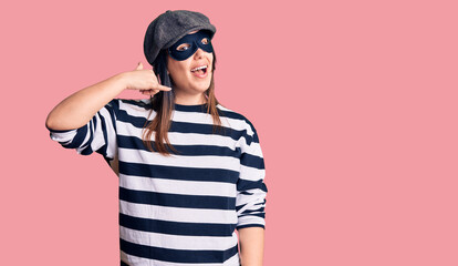 Young beautiful brunette woman wearing burglar mask smiling doing phone gesture with hand and fingers like talking on the telephone. communicating concepts.