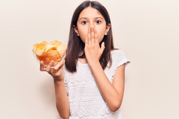 Beautiful child girl holding potato chip covering mouth with hand, shocked and afraid for mistake. surprised expression