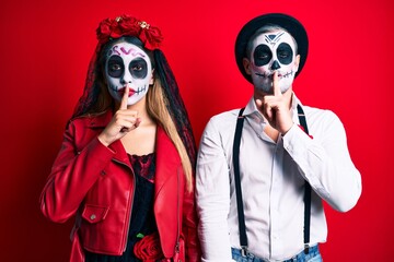 Couple wearing day of the dead costume over red asking to be quiet with finger on lips. silence and secret concept.