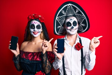 Couple wearing day of the dead costume holding smartphone showing screen smiling happy pointing with hand and finger to the side