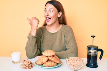 Young brunette woman sitting on the table having breakfast pointing thumb up to the side smiling happy with open mouth