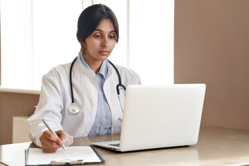 Fototapeta na wymiar Indian ethnic female doctor gp wearing white coat stethoscope writing watching online medical webinar seminar training or virtual meeting remote patient by video call working on laptop at workplace.