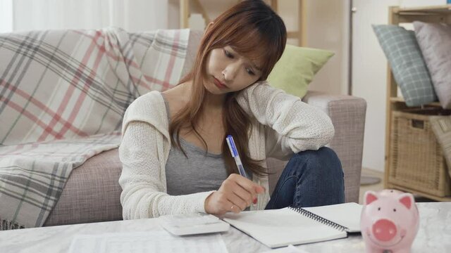 young woman tilting head pen tapping on notebook is considering budget control in tax season. asian girl sitting with piggy bank and calculator feels unhappy unable to buy clothes after paying taxes.