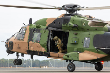 Camouflaged military helicopter touching down with a crewman looking on from the rear cabin.
