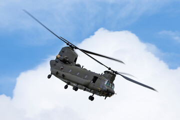 Large twin rotor military helicopter maneuvering through the blue sky in front of a large cloud formation.
