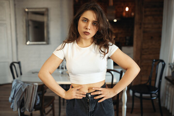 Portrait of unhappy grumpy young Caucasian woman in jeans and white top holding hands on her waist posing indoors, has to stay home, having displeased facial expression. People and lifestyle