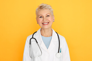 Isolated shot of confident experienced middle aged female doctor with short blonde haircut looking at camera with happy smile, wearing white medical coat and stethoscope around her neck - Powered by Adobe