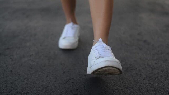 Closeup of woman's white sneakers walking towards camera on cracked asphalt