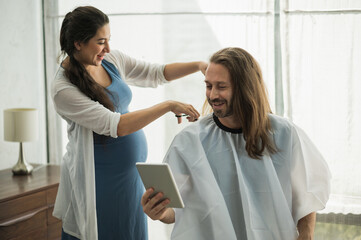 The pregnant woman is cutting her husband. Bearded man getting haircut by his wife at home new normal concept.
