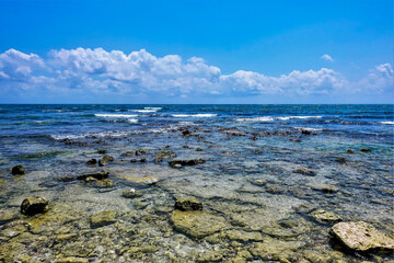 Caribbean Sea on a sunny summer day. There is clear water near the shore, flat stones are visible at the bottom. In the distance, turquoise waves with white foam. Azure sky with picturesque clouds. 