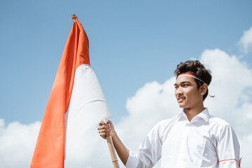 young man wearing a white shirt and red and white attributes holding stick with Indonesian flag against the background of the sky
