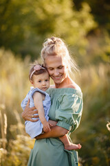 Mother and little daughter walk in blooming summer meadow. Mom loves her child. Toddler girl in Mom's hands. Happy family in beautiful summer day in flowering grass. Tender relations. Cotton outfit