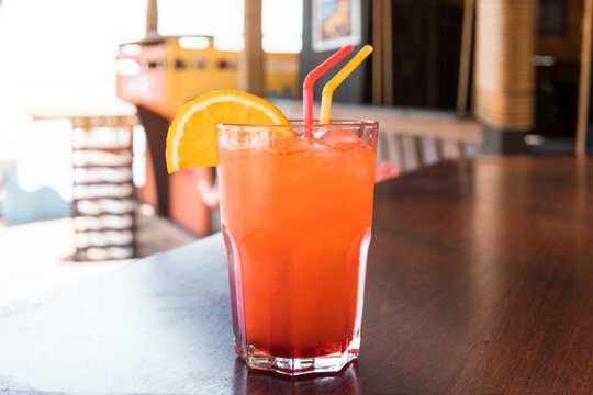 A Glass Of Red Juice On A Wooden Table. Closeup Ocktail Of Sweet Bourbon Whiskey Drink Garnished By Orange Peel On Table Diner Booth With Blurry Bokeh Restaurant Background