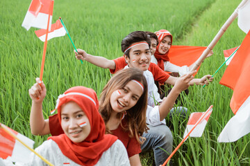 Asian young men and women hold and wave the Indonesian flag while wearing red and white attributes in the rice fields against a background of natural scenery