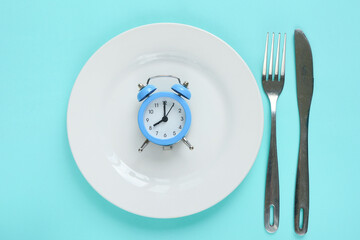 Blue alarm clock, fork, knife and empty plate on colored paper background. Intermittent fasting...