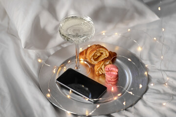 hygge and lifestyle concept - glass of champagne wine, smartphone, croissants, macaroons and garland lights in bed at home