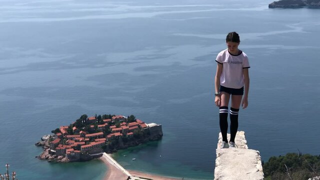 Young beautiful girl stands on edge of viewpoint of Sava Monaster. Scenic seashore view of Adriatic sea and Sveti Stefan island