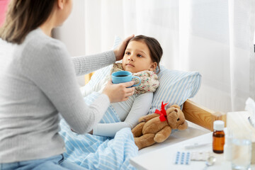 family, health and people concept - mother giving mug with hot tea to sick little daughter lying in bed at home