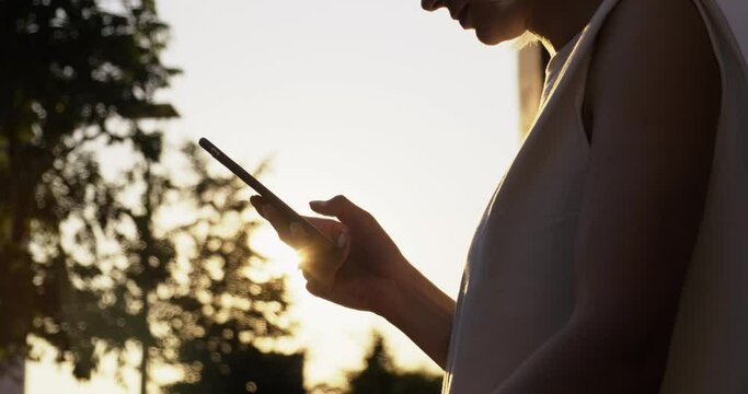 Close-up of Female Hands with Phone. Girl Standing on Street at Sunset. Writes Message. Chatting. Communicates with Friends and Followers. Silhouette. Summer. City Street in Europe. Sun Glare. Evening