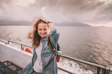 Happy curly young woman traveler on a ship at the sea against the backdrop of a beautiful landscape. Windy weather and flying hair, adventure travel concept