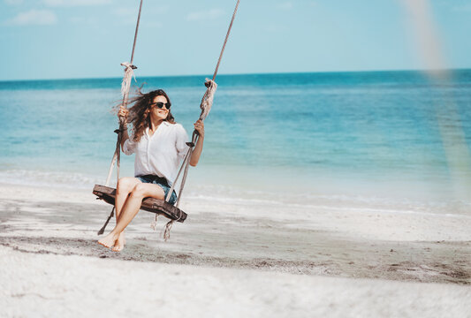 Young beautiful curly woman girl swinging and having fun on a swing on a tropical beach vacation and travel concept.