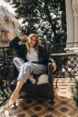 Stylish fashionable blonde woman with smoky eye makeup, in jeans, white T-shirt and black leather jacket on the balcony in an armchair. Spring autumn fashion concept. Soft selective focus.
