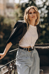Stylish fashionable blonde woman with smoky eye makeup, in jeans, white t-shirt and black leather jacket on the balcony in the city. Spring autumn fashion concept. Soft selective focus.