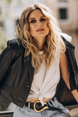 Fototapeta na wymiar Stylish fashionable blonde woman with smoky eye makeup, in jeans, white t-shirt and black leather jacket on the balcony in the city. Spring autumn fashion concept. Soft selective focus.