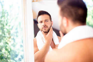 Handsome young man standing in front of bathroom mirror and looking his face