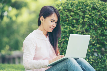 Asian woman sitting green park using laptop computer. Woman working on laptop happy entrepreneur business using notebook with hands typing on keyboard home office during coronavirus quarantine period