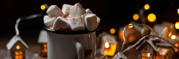 Winter hot drink: hot chocolate with marshmallows, cinnamon and anise. Christmas lights in the shape of houses as decor. Decorative snow, dark wooden background. Festive mood, cozy home atmosphere