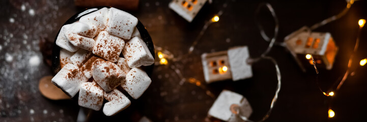 Winter hot drink: hot chocolate with marshmallows, cinnamon and anise. Christmas lights in the shape of houses. Decorative snow, dark black background. Festive mood, cozy atmosphere. Flat lay top view
