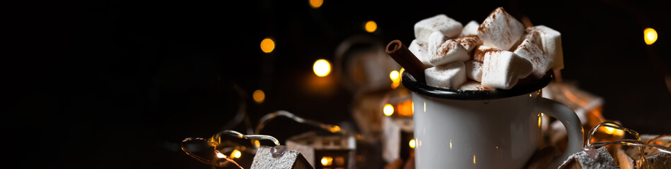 Winter hot drink: hot chocolate with marshmallows, cinnamon and anise. Christmas lights in the shape of houses as decor. Decorative snow, dark wooden background. Festive mood, cozy home atmosphere