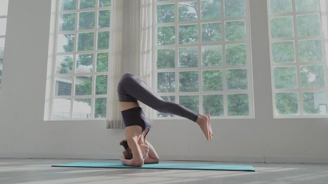wide shot: Young Asian sporty woman  practicing yoga on mat in headstand pose upside down balancing body at fitness studio. healthy and wellness lifestyle
