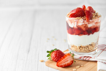 Homemade layered dessert with fresh strawberries, cream cheese or yogurt, granola and strawberry jam in glass on white wood background. Healthy organic breakfast or snack concept. Selective focus.