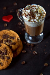 Close up of a glass cup of coffee with whipped cream and chocolate on it, chocolate chip cookies and roasted coffee beans on dark background. Concept of ready to eat food, tasty snack. Selective focus