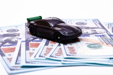 Black toy car on a heap of dollar bills on a white and black background. A sports car with a green spoiler, a wing on dollars in a close-up top and side view. Rear and front car. Selling, buying