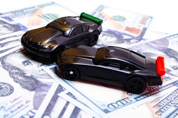 Two black toy cars on a heap of dollar bills on a white and black background. Two sports cars on...