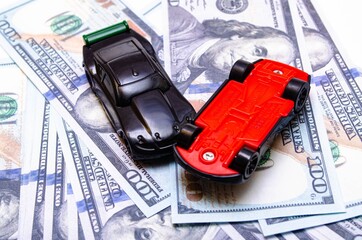 Two black toy cars on a heap of dollar bills on a white and black background. Two sports cars on...