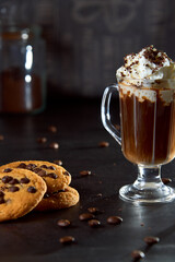 Close up of a glass cup of coffee with whipped cream and chocolate on it, chocolate chip cookies and roasted coffee beans on dark background. Concept of ready to eat food, tasty snack. Selective focus