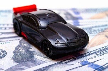 Black toy car on a heap of dollar bills on a white and black background. A sports car with a red...