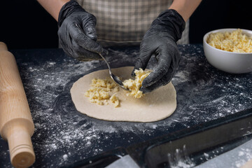 Woman in checkered apron and black gloves spread the filling on the dough. Process of making bakery. Adjarian Khachapuri Recipe – Georgian cheese bread.
