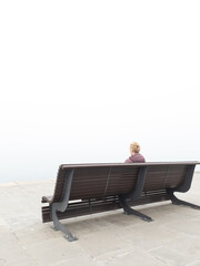 Woman in despair sitting on a lonely bench and looking at grey distant horizon on a ocean foggy day.