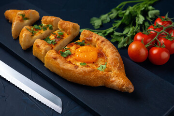 Sliced Adjarian Khachapuri – Georgian cheese bread decorated with tomatoes and parsley on dark blue textured background. Baked pie with cheese and egg yolk filling.