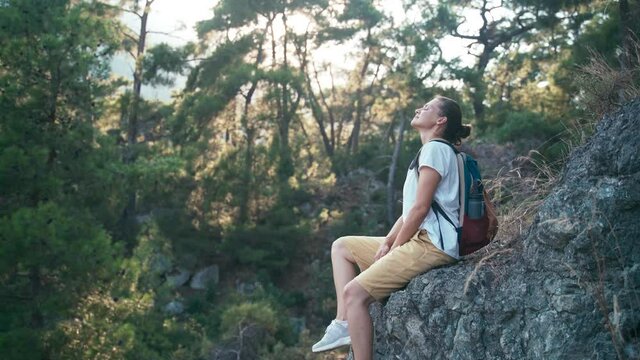 Young woman traveler with a backpack sitting on a rocky mountainside and enjoying fresh mountain air while resting.