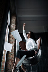 Business shot of a business woman in a white shirt sitting in a metal aviator chair and throwing...