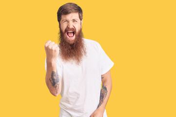 Handsome young red head man with long beard wearing casual white tshirt angry and mad raising fist...