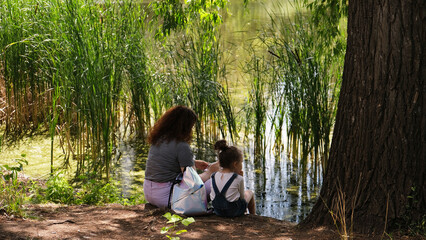 mother and daughter are sitting on the shore of a pond overgrown with reeds