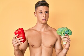 Young hispanic boy shirtless holding broccoli and red pepper clueless and confused expression. doubt concept.